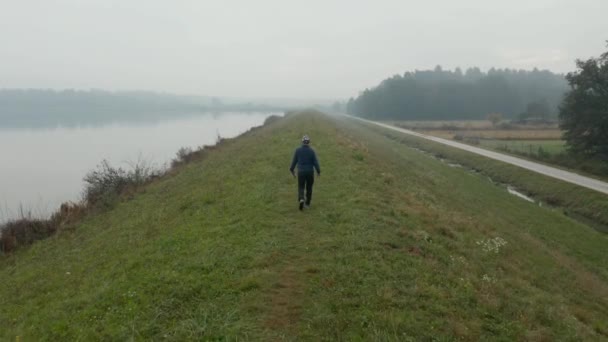 Droneskudd forfølger en mann som går på en gressaktig høyde med utsikt over en reflekterende innsjø. – stockvideo
