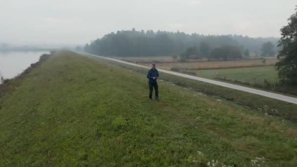 Чоловік, який керує безпілотником на трав'яному пагорбі з туманним озером на задньому плані, повітря — стокове відео