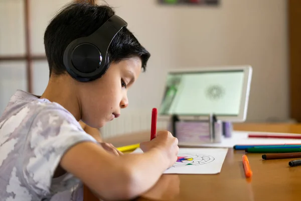 Sevimli bir çocuğun renkli kalemleriyle mandalayı boyayıp evdeki kulaklıklarla tabletinde özel ders izlerken çekilmesi.