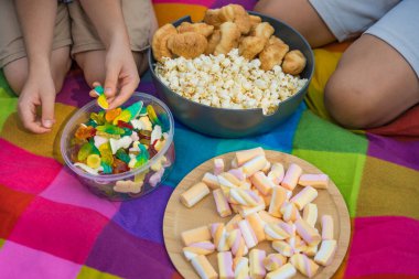 İki çocuk gökkuşağı battaniyesinde patlamış mısır, kruvasan, şekerleme ve şekerleme kaselerinde oturarak yaz pikniğinin tadını çıkarıyor..