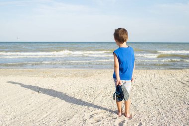 Plajda denize bakarak genç çocuk. Küçük çocuk suyun kenarında plajda yürüyüş arkadan görünüm