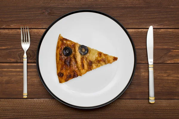 A piece of pizza on white plate with a knife and fork on wooden background. Slice of tasty pizza on the plate. Top view