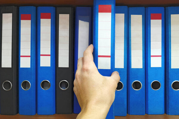 Office worker taking a folder in the archive: database, administration and file management concept. Closeup hand selecting big folder from the shelves with office files