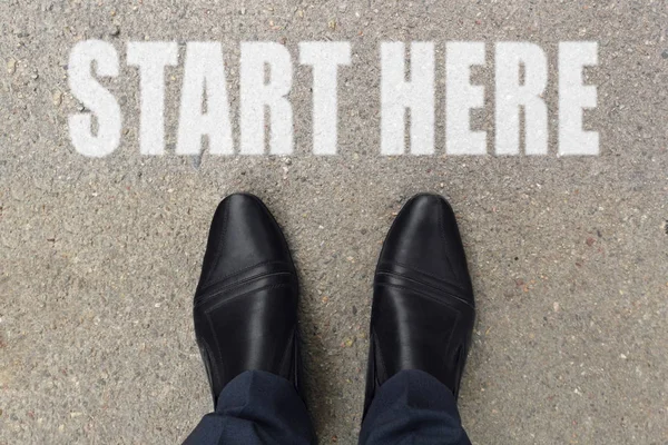 Businessman is looking down at his feet on a concrete floor with START HERE letters painted on the surface. Top view image of person on road with the text start here