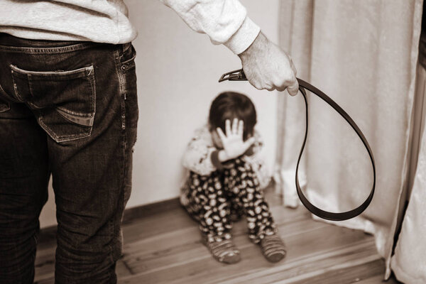 Aggressive parent with belt and frightened child in corner. Domestic violence. Abused child, aggression in the family