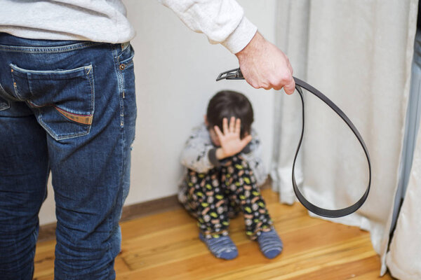 Aggressive parent with belt and frightened child in corner. Domestic violence. Abused child, aggression in the family