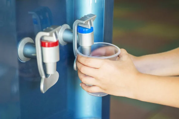 Mano del niño sosteniendo el vaso con agua del enfriador de agua. Pequeño niño presiona el agua potable — Foto de Stock