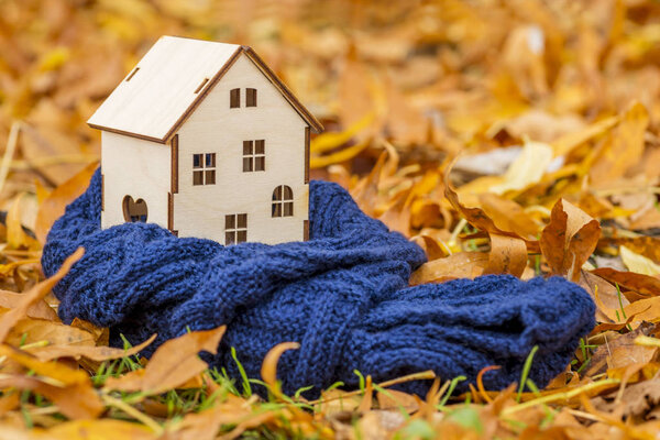 Игрушечный деревянный дом завернут в теплый шарф на фоне осенних листьев. Концепция теплого дома
