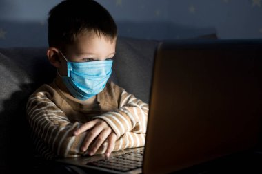 Tıbbi maskeli çocuk deftere bakıyor ve akşam yeni bilgileri alıyor, karantina sırasında online eğitim alıyor..