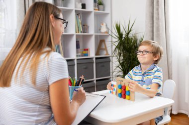 Genç bir çocuk evdeki bir öğrenme seansı sırasında öğretmeniyle etkileşime girer. Çocuk renkli bir eğitim oyuncağı tutarken, öğretmen notlar alıyor ve olumlu bir öğrenme ortamı sağlıyor..