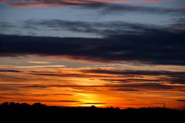 Sıcak turuncu, kırmızı ve mavi tonlarda bulutların oluşturduğu gün batımının çarpıcı geniş açılı görüntüsü. Bir ağaç hattının koyu silueti çerçevenin altında görünür..