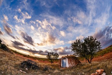 Beyaz kapılı taştan bir barakanın geniş, manzaralı bir görüntüsü, birkaç ağaçla birlikte kuru, çimenli bir arazide geçiyor. Yukarıdaki gökyüzü mavi ve altın tonlarında yumuşak, ince bulutlarla dolu..