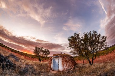 Yıpranmış kapısı olan küçük yuvarlak, yuvarlak bir taş kulübenin biraz yüksek, geniş açılı görüntüsü. Kulübe, her iki tarafında iki ağaç olan kuru, yabani otlar ve kırmızımsı toprakların olduğu bir arazide duruyor. Gökyüzü bulutlarla ve zengin, sıcak bir günbatımıyla dolu.