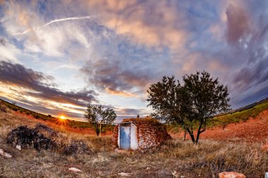 Geniş açılı bir manzara, günbatımında sakin bir sahneyi yakalıyor. Merkezde, taştan yapılmış küçük, dairesel bir kulübe iki ağacın arasında duruyor. Batan güneş ufukta beliriyor, bulutlara ve kuru, çimenli alana sıcak bir parıltı saçıyor. Vibratör.