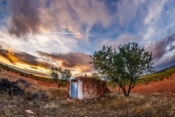 Düşük açılı, geniş açılı, kurumuş taşlardan yapılmış küçük, geleneksel bir kulübe. İki ağaç yapının etrafını sarıyor, koyu yaprakları renkli gökyüzüne zıtlık yaratıyor. Sahne, gün batımının sıcak, altın ışığıyla aydınlatılıyor, bir baloyla.