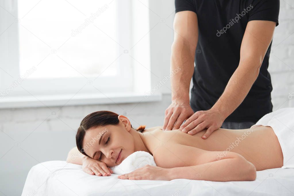 Young beautiful woman enjoying a back massage. Professional massage therapist is treating a female patient in apartment. Relaxation, beauty, body and face treatment concept. Home massage.
