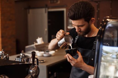 Genç, gülümseyen erkek barista cappuccino yapıyor. Kahve hazırlamak için süt dolduruyorum. Latte sanatı. Kafede sabah kahvesi. Kahve dükkanı konsepti.