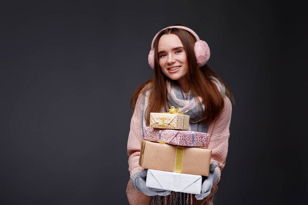 Young beautiful smiling girl in knitting pullover and pink fluffy earmuffs holding gift boxes on a gray background. Xmas model with long straight hair. Winter holidays, Christmas, New Year concept. 