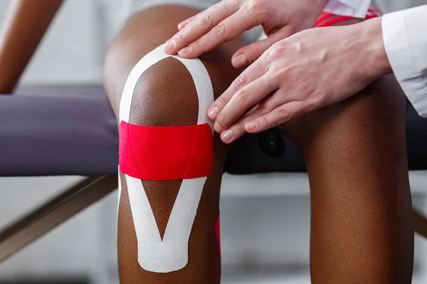 Physiotherapist applying kinesiology tape to patient knee.Therapist treating young female African American athlete. Kinesiology taping. Post traumatic rehabilitation,sport physical therapy