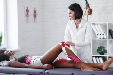 Fizyoterapist hastanın dizine kineziyoloji bandı uyguluyor. Genç Afrikalı Amerikalı atleti tedavi eden terapist. Kineziyoloji bandı. Travma sonrası rehabilitasyon, spor fizyoterapisi