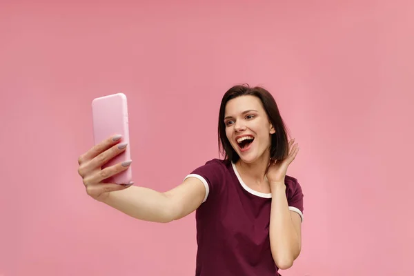 Funny young brunette woman making selfie. Smiling girl wearing t-shirt holding pink smartphone, making faces on camera, posing for selfie isolated on pink background — Stock Photo, Image