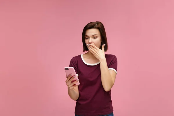 Surpised young brunette woman holding pink smartphone, smiling and expressing positivity. Happy girl got shocking positive news. Copy space. Young people working with mobile devices — Stock Photo, Image