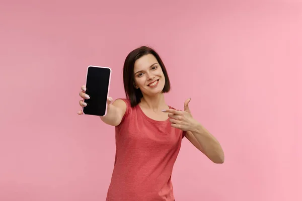 Funny brunette woman showing blank screen mobile phone on camera. Young smiling girl demonstrating smartphone display isolated over pink background. Copy space. App for your business — Stock Photo, Image