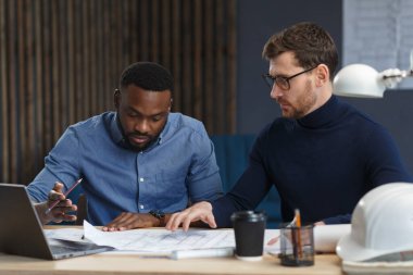 Çok ırklı mühendis ekibi mavi baskılar ve mimar ekipmanlarıyla birlikte çalışıyor, taslak çiziyorlar, tartışıyorlar ve inşaat projesini planlıyorlar. Ortaklar iş yerinde tartışır. İş inşa kavramı