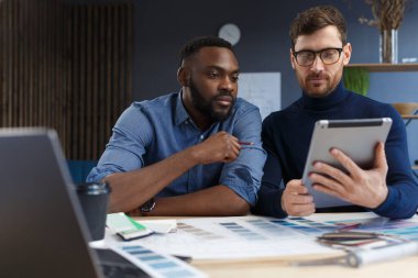 Çok ırklı mühendis ekibi tablet bilgisayarda çalışıyor. Mimarlar bina renklerini renk örnekleri, çizim ve inşaat projeleri kullanarak seçerler. İş yerinde tartışan ortaklar