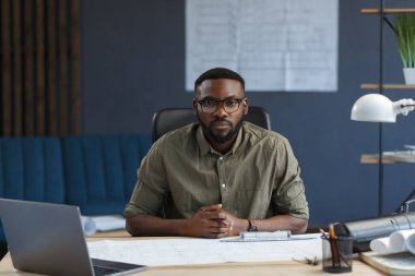 Ofiste dizüstü bilgisayarla çalışan Afro-Amerikalı bir mimar. İş yerinde oturan, gözlüklü, siyah, yakışıklı, sakallı bir adamın iş portresi. Kendinden emin iş adamı başarılı oldu.