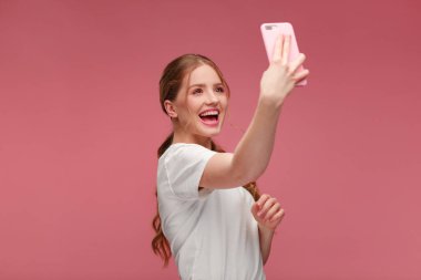 Komik kızıl saçlı kadın selfie çekiyor. Beyaz tişört giyen, elinde pembe akıllı telefon tutan gülümseyen kız, kamerada yüz ifadeleri yapan, pembe arka planda izole edilmiş selfie için poz veren kız.