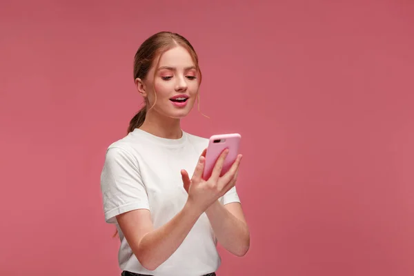 Lustige junge rothaarige Frau mit rosa Smartphone und Lächeln. Glückliches Mädchen mit Handy-Apps, SMS, Surfen im Internet, Blick auf das Smartphone. Junge Leute, die mit mobilen Geräten arbeiten. — Stockfoto