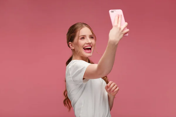 Funny young redhead woman making selfie. Smiling girl wearing white t-shirt holding pink smartphone, making faces on camera, posing for selfie isolated on pink background — Stock Photo, Image
