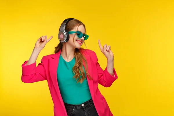 Müzik dinlemenin tadını çıkar. Kulaklıklı genç kızıl kadın müzik dinliyor. Kulaklıklı, pembe ceketli, dans eden ve sarı arka planda şarkı söyleyen komik kız. Rahatlama ve stres yönetimi