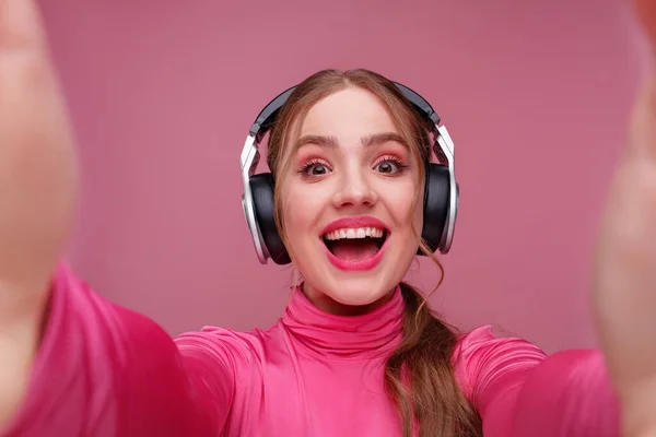 Retrato de cerca de una joven pelirroja sonriente haciendo selfie usando dos manos. Chica sonriente divertida con auriculares que dan un regalo aislado en el fondo rosa. Copiar espacio —  Fotos de Stock
