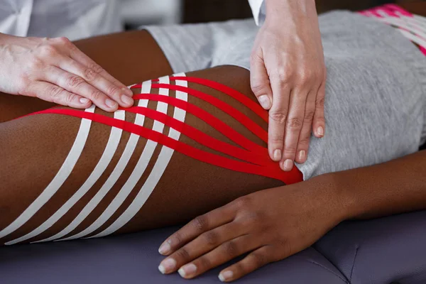 キニソロジーテープ。理学療法士は患者の腰に運動テープを適用します.若い女性アフリカ系アメリカ人選手を治療セラピスト.外傷後リハビリテーション,脂肪減少,スポーツ理学療法 — ストック写真