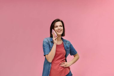 Mobil cihazlarla çalışan gençler. Tişört ve kot pantolon giyen, cep telefonuyla konuşan pembe arka planda gizlenmiş gülümseyen güzel esmer kadın portresi. Boşluğu kopyala