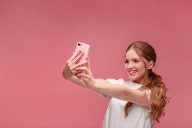 Komik kızıl saçlı kadın selfie çekiyor. Beyaz tişört giyen, elinde pembe akıllı telefon tutan gülümseyen kız, kamerada yüz ifadeleri yapan, pembe arka planda izole edilmiş selfie için poz veren kız.