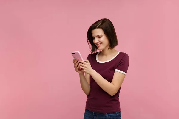 Funny young brunette woman holding pink smartphone and smiling.Happy girl using mobile phone apps, texting message, browsing internet, looking at smartphone. Young people working with mobile devices — Stock Photo, Image