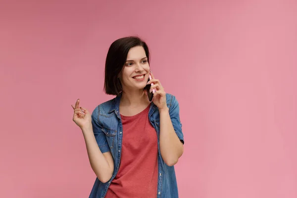 Young people working with mobile devices. Portrait of cute happy brunette woman wearing t-shirt and jeans shirt talking on mobile phone and smiling isolated over pink background. Copy space