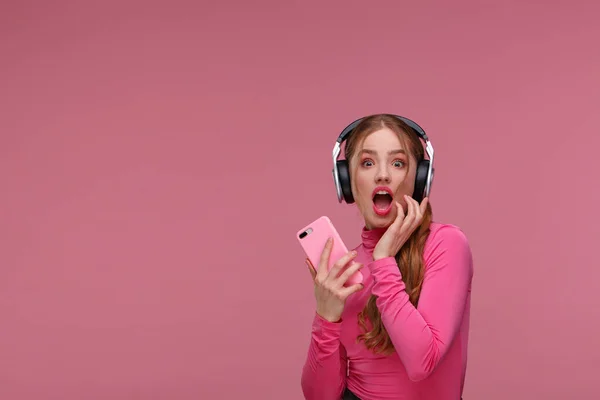 Funny surprised redhead girl holding mobile phone waiting for messages and congratulations from friends. Young smiling woman wearing earphones holding smartphone over pink background. Copy space — Stock Photo, Image