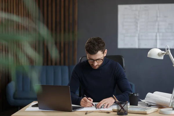 Arquitecto trabajando en oficina con laptop. Ingeniero piensa en el plan arquitectónico, la búsqueda de nuevas ideas para el proyecto de construcción. Retrato del guapo barbudo sentado en el lugar de trabajo. Concepto empresarial —  Fotos de Stock