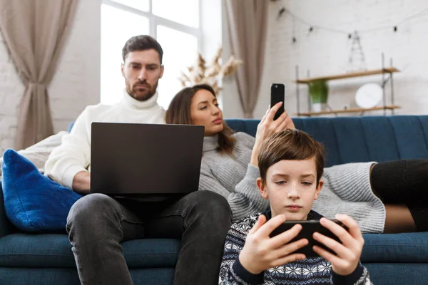 Madre, padre e hijo usando laptop y teléfonos móviles en casa. Miembros de la familia ignorándose unos a otros y hablando en vivo. Gadget influencia en las relaciones familiares. Tecnologías modernas y concepto de adicción —  Fotos de Stock