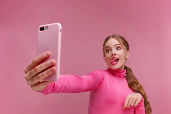 Joven pelirroja divertida haciendo selfie. Chica sonriente con blusa rosa sosteniendo teléfono inteligente rosa, haciendo caras en la cámara, posando para selfie aislado sobre fondo rosa —  Fotos de Stock