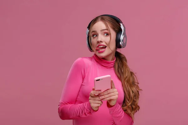 Chica pelirroja divertida sosteniendo el teléfono móvil esperando mensajes y felicitaciones de amigos. Joven mujer sonriente con auriculares sosteniendo teléfono inteligente sobre fondo rosa. Copiar espacio —  Fotos de Stock