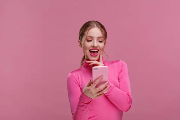 Il miglior giorno di sempre. Giovane donna dai capelli rossi sorpresa con uno smartphone rosa, sorridente ed espressiva positività. La ragazza felice ha ricevuto notizie positive scioccanti. Ricevuto. Giovani che lavorano con dispositivi mobili — Foto Stock