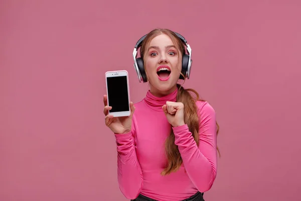 Chica pelirroja divertida mostrando la pantalla en blanco del teléfono móvil en la cámara. Mujer sonriente joven con auriculares que demuestran la pantalla del teléfono inteligente aislado sobre fondo rosa. Copiar espacio. Aplicación para su negocio. —  Fotos de Stock