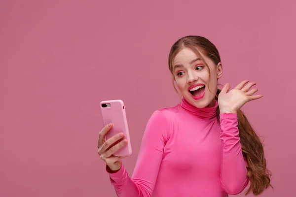 Найкращий день у світі. Здивована молода руда жінка тримає рожевий смартфон, посміхаючись і висловлюючи позитивізм. Щаслива дівчина отримала шокуючі позитивні новини. Копіювати простір. Молоді люди, що працюють з мобільними пристроями — стокове фото