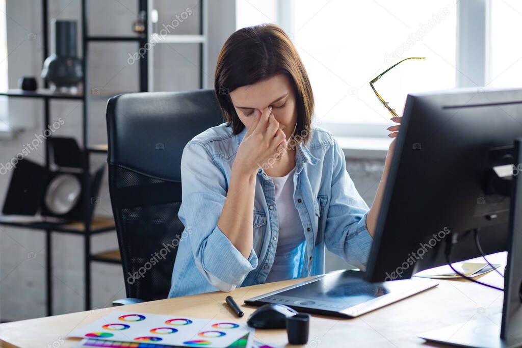 Portrait of tired graphic designer working overtime in office. Stressed worker have eye strain symptoms. Retoucher workplace in photo studio. Burnout and overwork concept. Creative agency