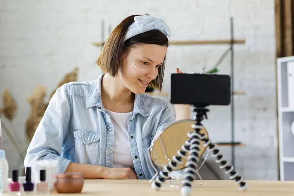 Young female blogger recording a tutorial video for her beauty blog about cosmetics.Vlogger testing face cream and broadcast live video to social network at home.Blogging, videoblog, skincare concept